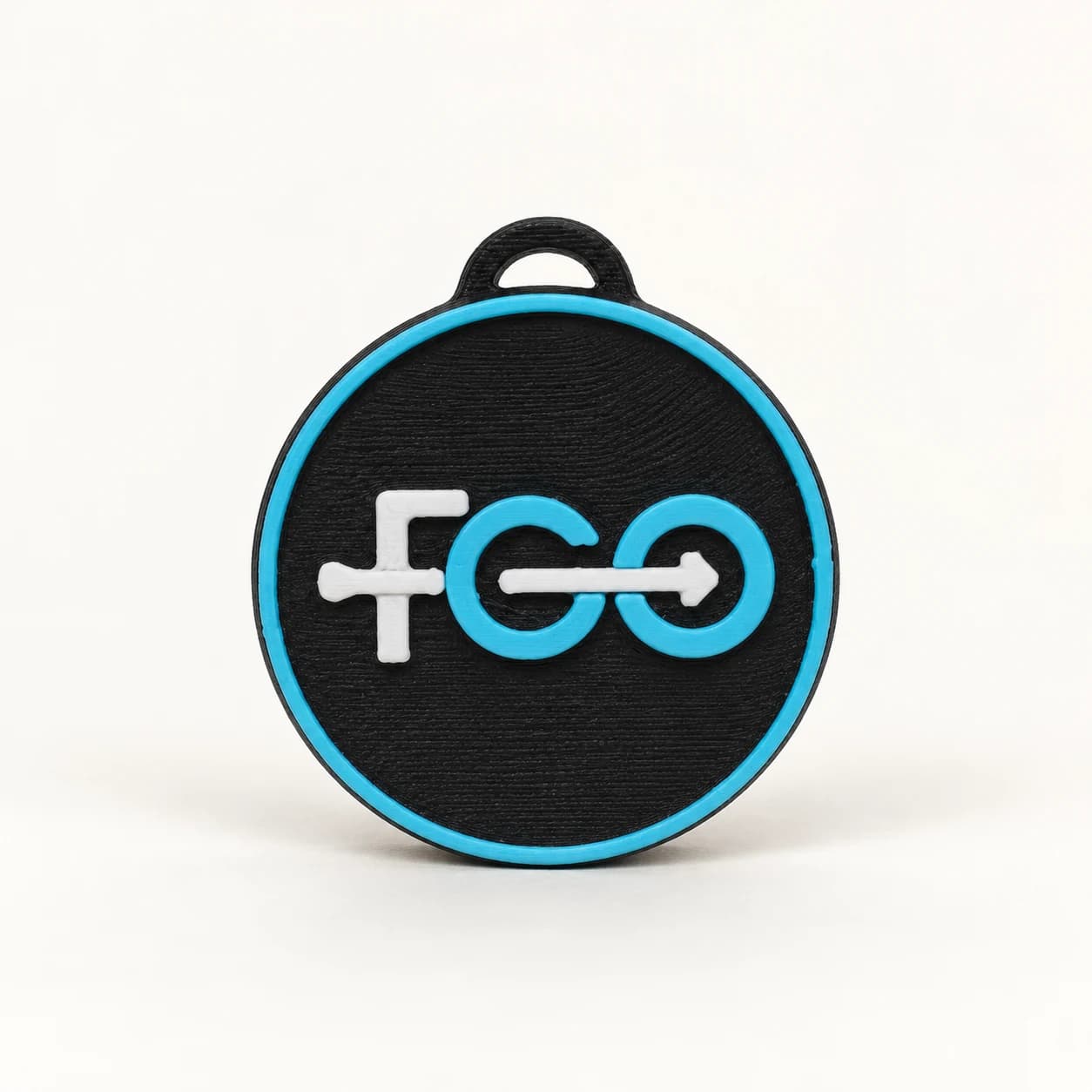 Round black FGO keyring with a white and turquoise logo, turquoise border and black loop, in a studio image.